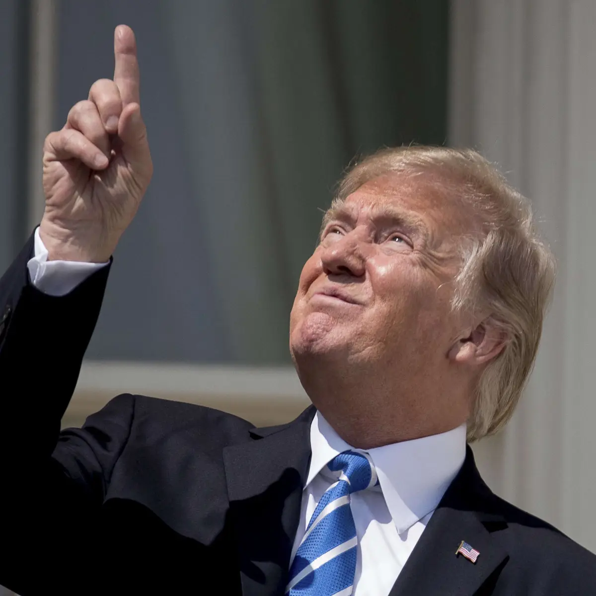 a man in a dark suit and blue tie with an american flag pin on his lapel looks upward with a smug expression while pointing…