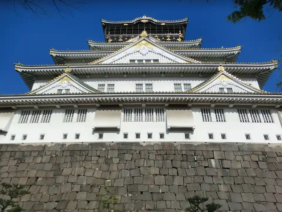 a low-angle photograph of Osaka Castle under a clear blue sky, showing its white walls, golden roof trim, and stone base…