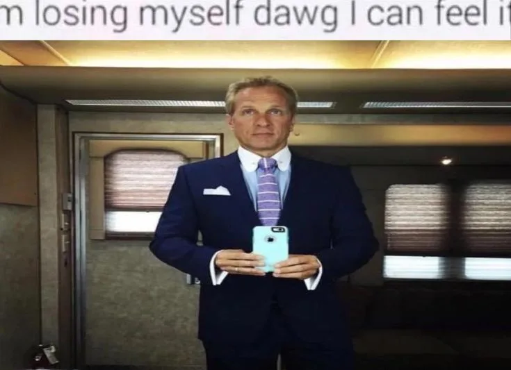 A man in a dark blue suit and striped purple tie stands in a luxurious train car, holding a light blue phone to take a…