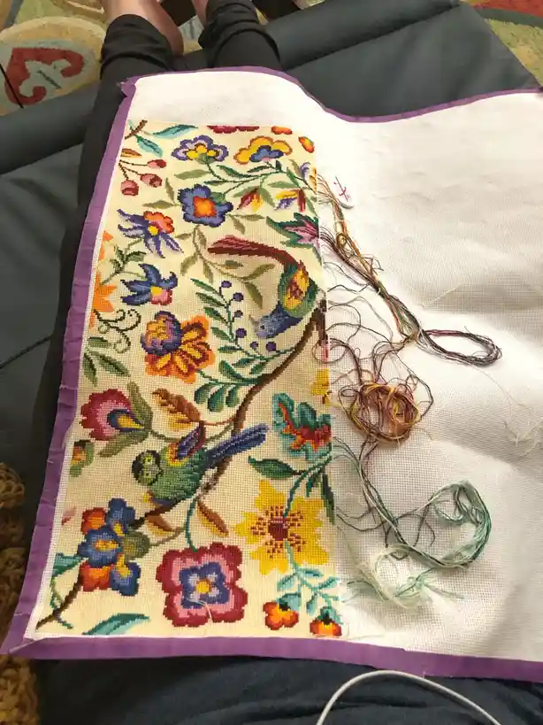 A person's legs are visible as they sit on a couch, holding a large cross-stitch project. The project shows a colorful…