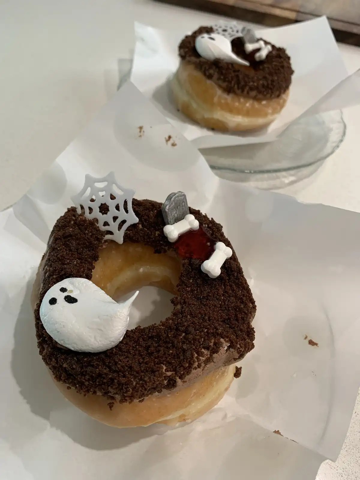 Two donuts are decorated with Halloween-themed toppings including ghost, spiderweb, tombstone, bones, and fake blood on a…