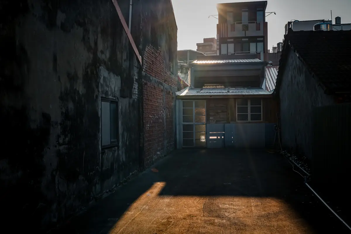 A narrow alleyway between weathered buildings with a single patch of sunlight illuminating the ground, creating a stark…
