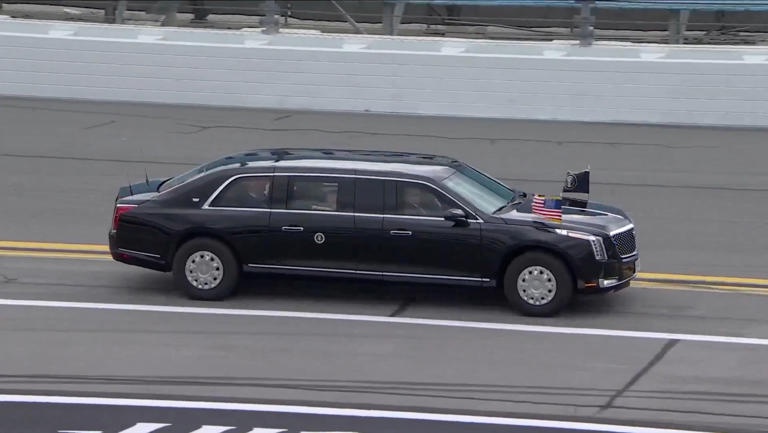 A black, elongated limousine, resembling a presidential motorcade vehicle, drives on a racetrack. Two small flags are…