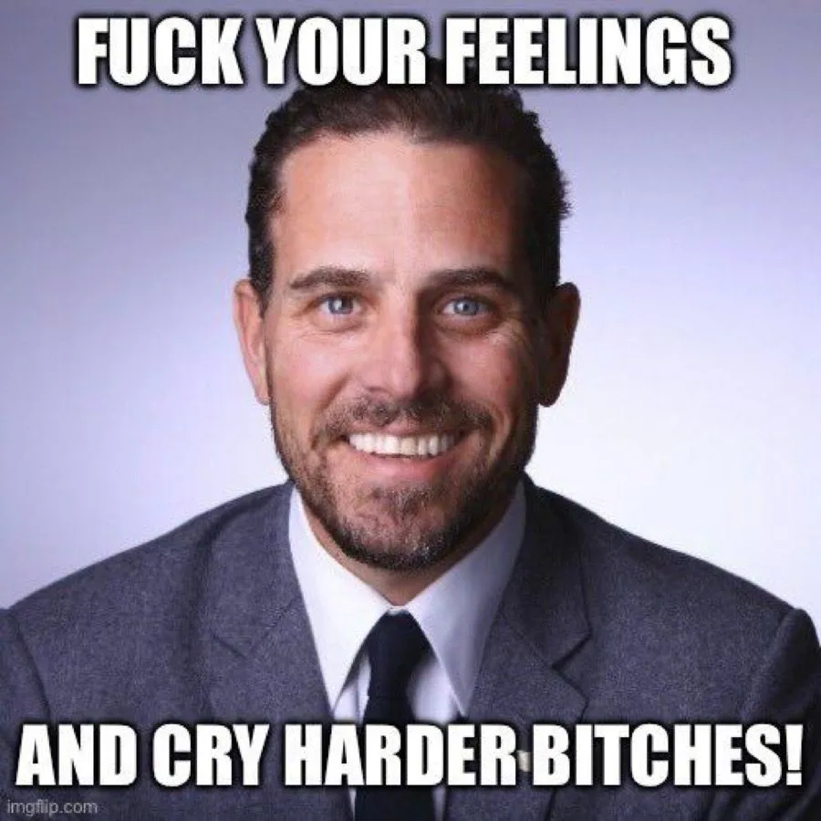 A man with blue eyes, a beard, and a wide, confident smile wears a gray suit, white shirt, and black tie against a plain…