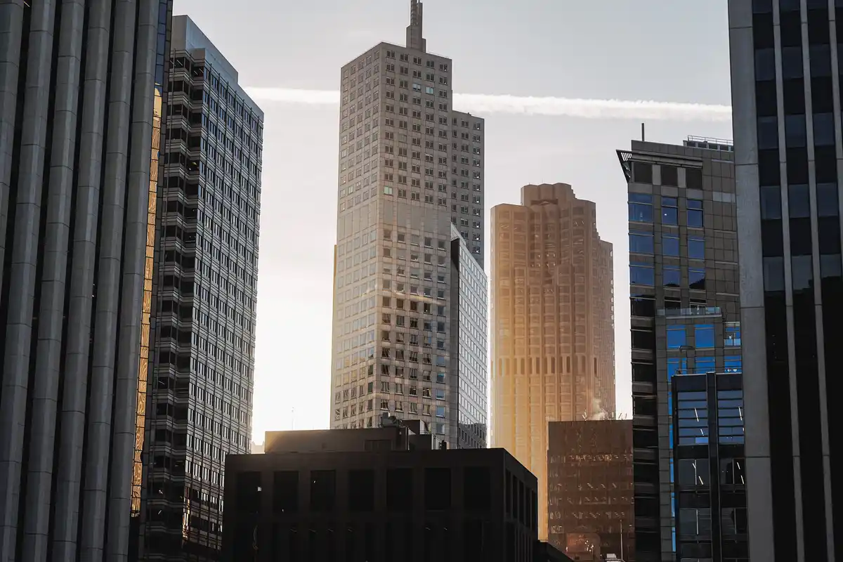 A cityscape of tall skyscrapers with sunlight hitting their glass facades, creating a warm golden glow. A thin white…