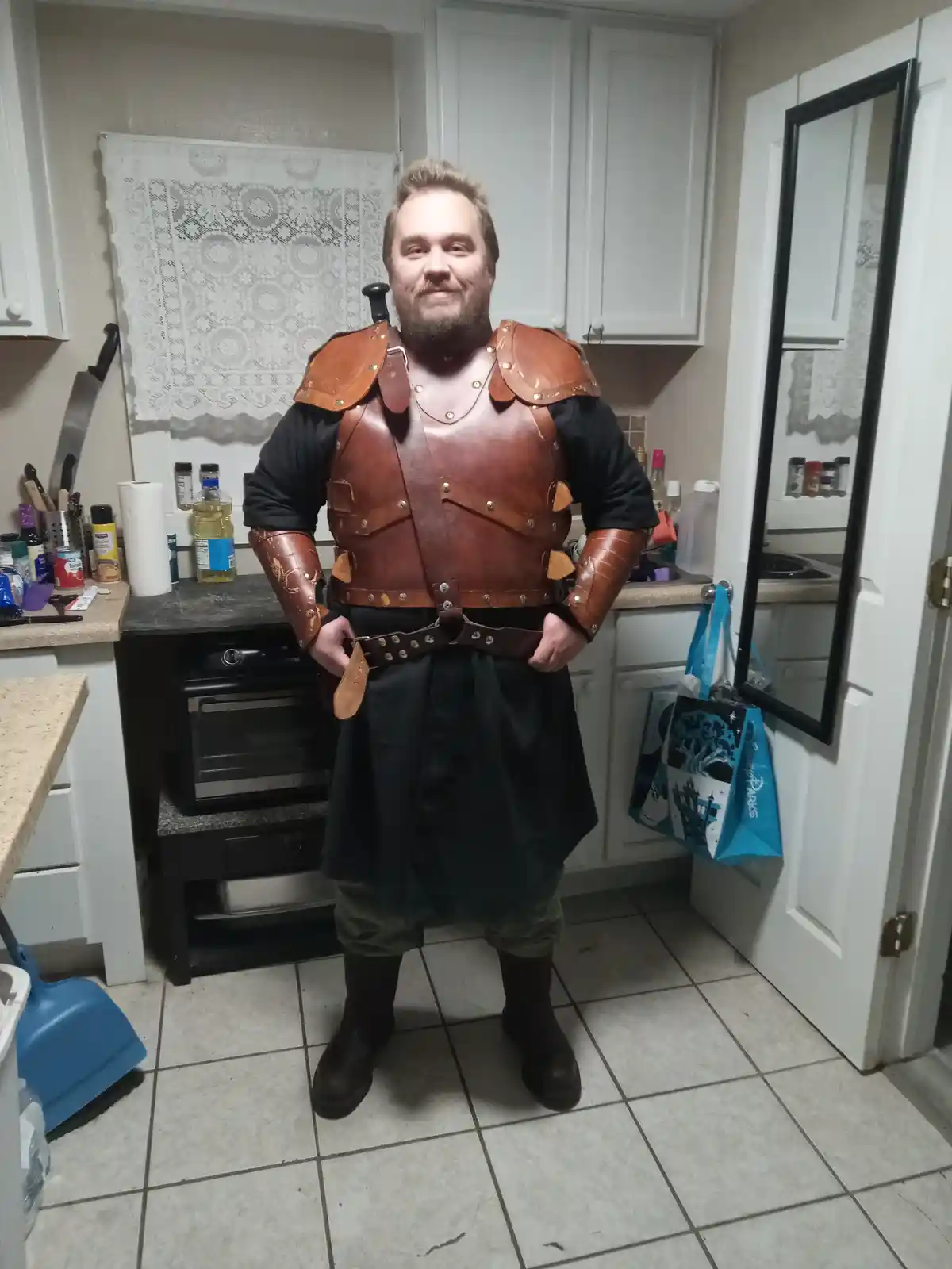 A man with a beard and short hair stands in a kitchen wearing full brown leather armor including a breastplate, shoulder…