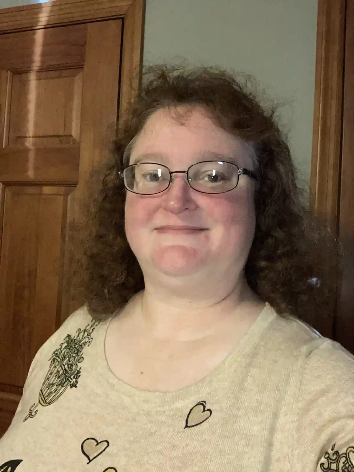 A person with curly brown hair and glasses smiles slightly while wearing a beige shirt with heart and herb designs, standing…