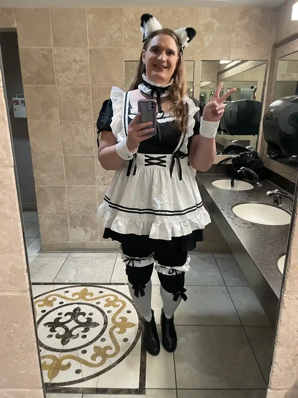 A person with long hair and panda ears takes a mirror selfie in a public restroom, wearing a black and white maid outfit…