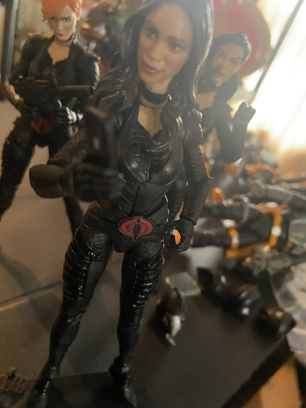 A close-up photo of a black-clad action figure, resembling a female Cobra Commander from G.I. Joe, holding a phone as if…