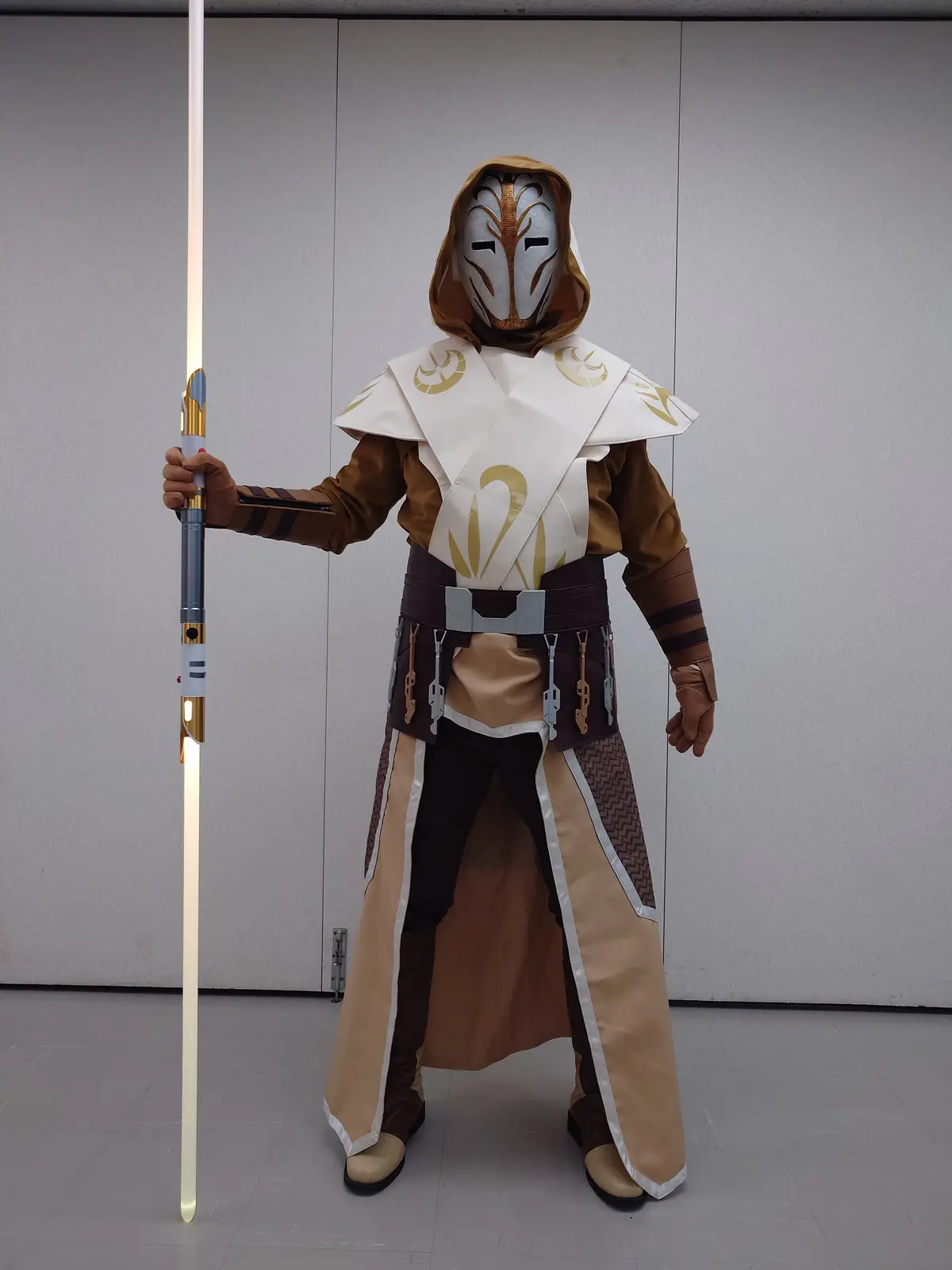A person in a full-body cosplay of a Jedi from Star Wars, wearing a distinctive white and brown hooded robe with gold…