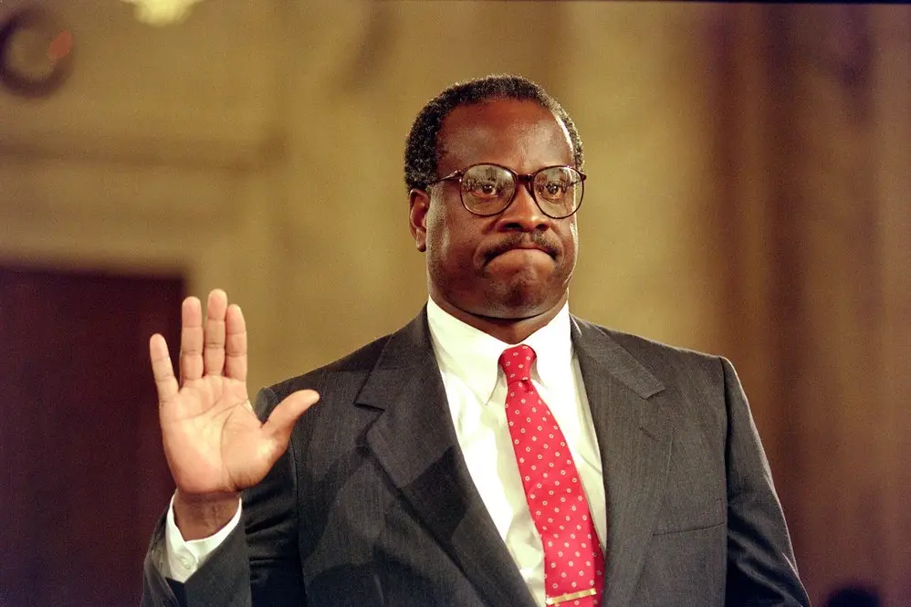 a man in a dark suit, white shirt, and red polka-dot tie raises his right hand while wearing thick-rimmed glasses, looking…