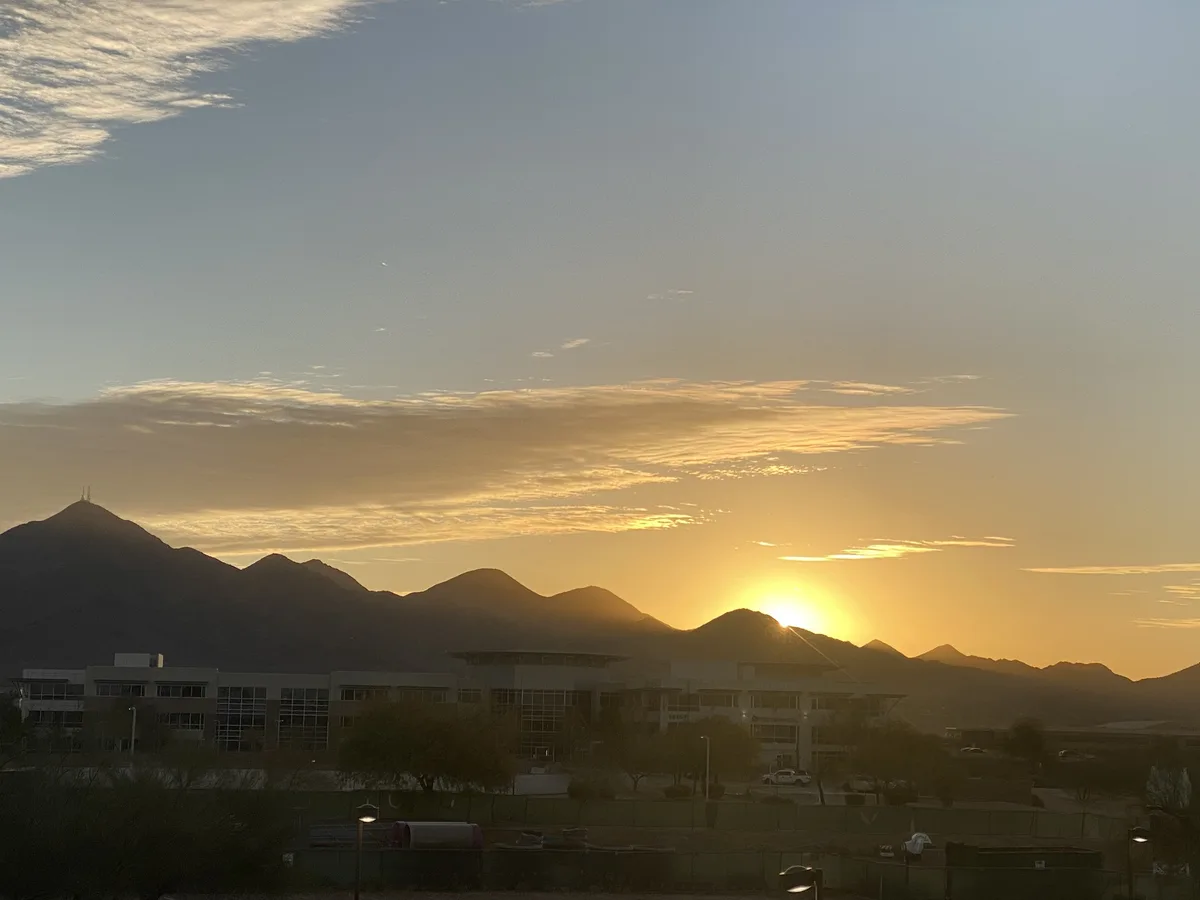 A scenic photo of a sunrise or sunset over a mountain range with a modern building in the foreground. The sun is low on the…