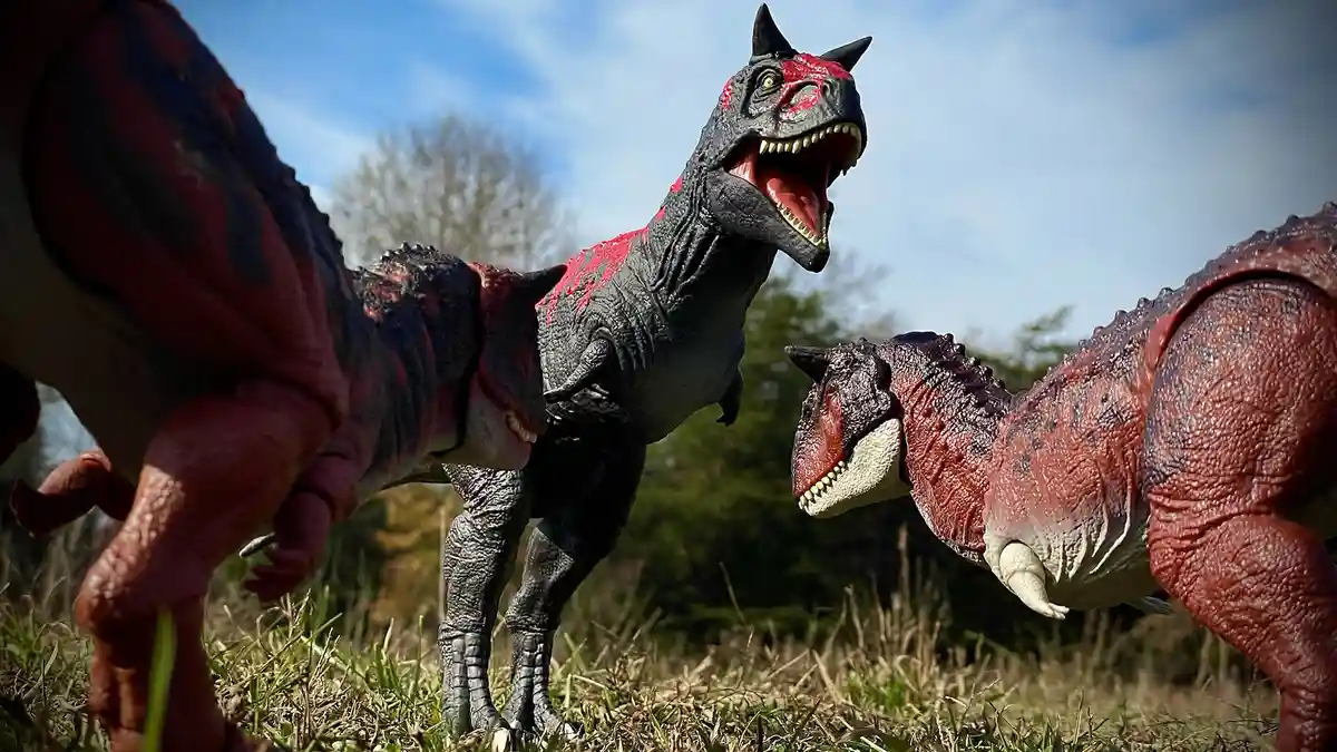 Three plastic dinosaur toys are posed in a grassy field under a blue sky, appearing to confront each other. The central…