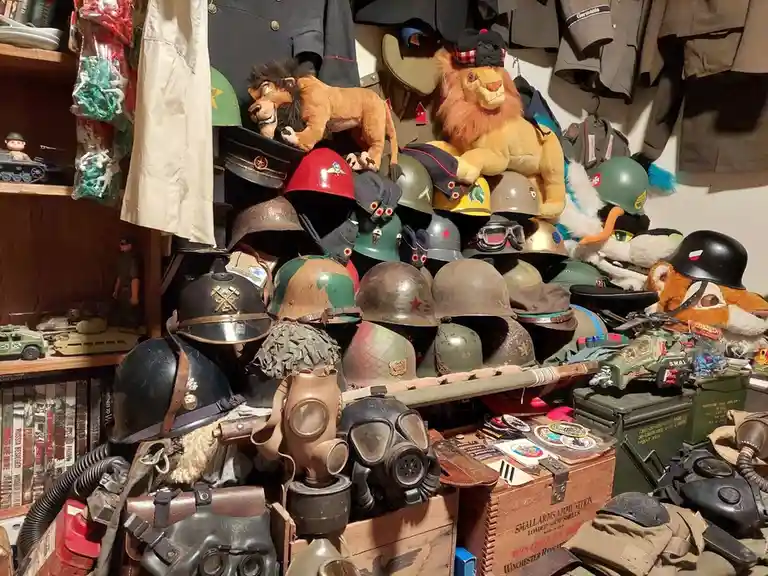 A cluttered collection of military helmets, gas masks, uniforms, and toy tanks is piled high, with several large plush lions…