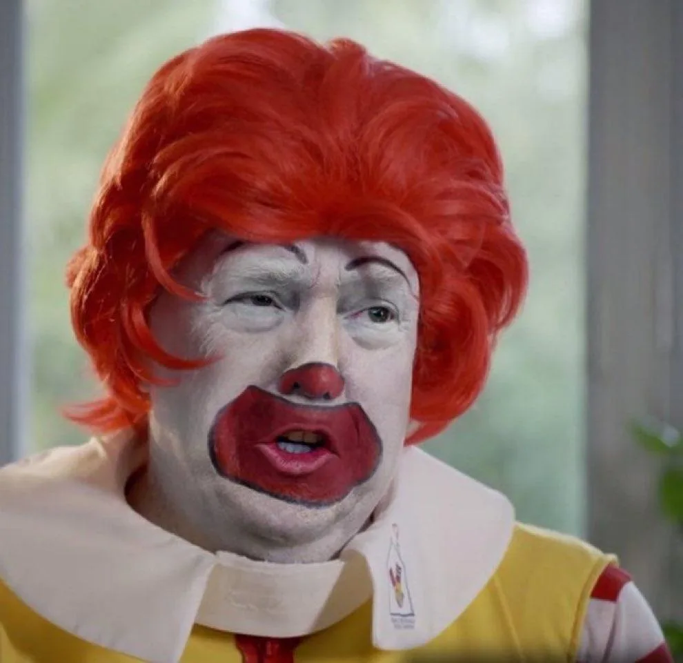 A person with a red wig, white face paint, red nose, and red lips resembling Ronald McDonald, wearing a yellow and white…