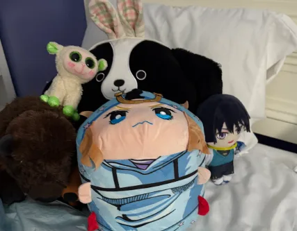 A collection of plush toys arranged on a bed, including a large central plushie of a character with a sad expression…