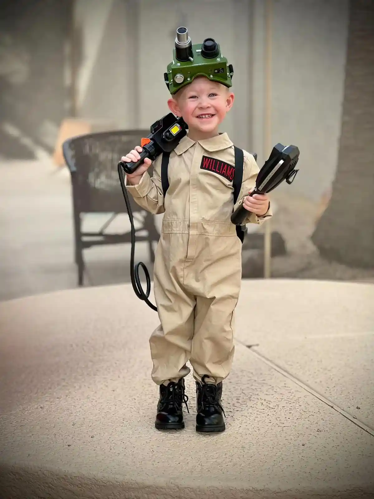 A young child dressed as a Ghostbuster, wearing a tan jumpsuit with 'WILLIAMS' on the chest, a green proton pack helmet, and…