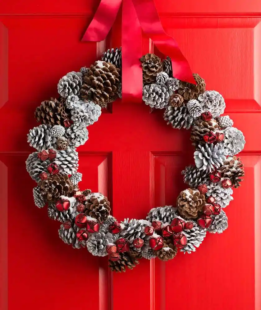 A festive holiday wreath made of brown and silver pinecones, adorned with small red jingle bells and tied with a red ribbon…