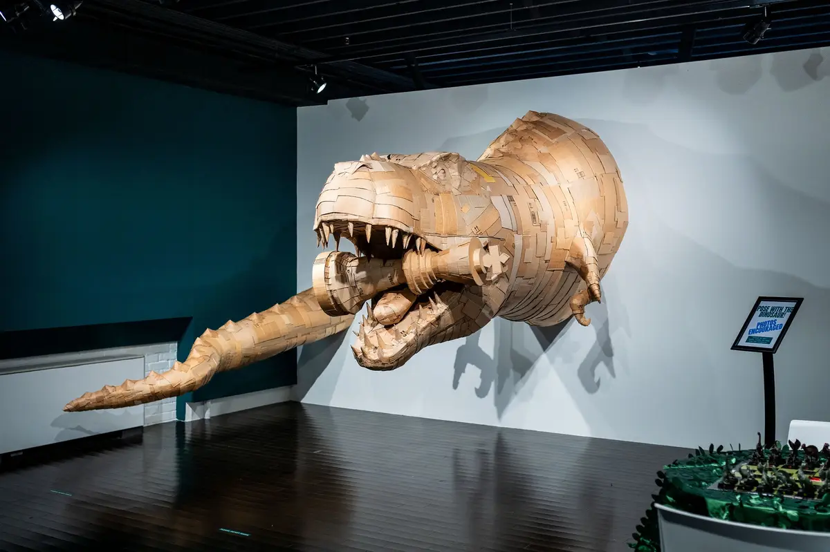 A large, cardboard sculpture of a Tyrannosaurus Rex is mounted on a wall in a museum-like setting, its mouth open and jaws…