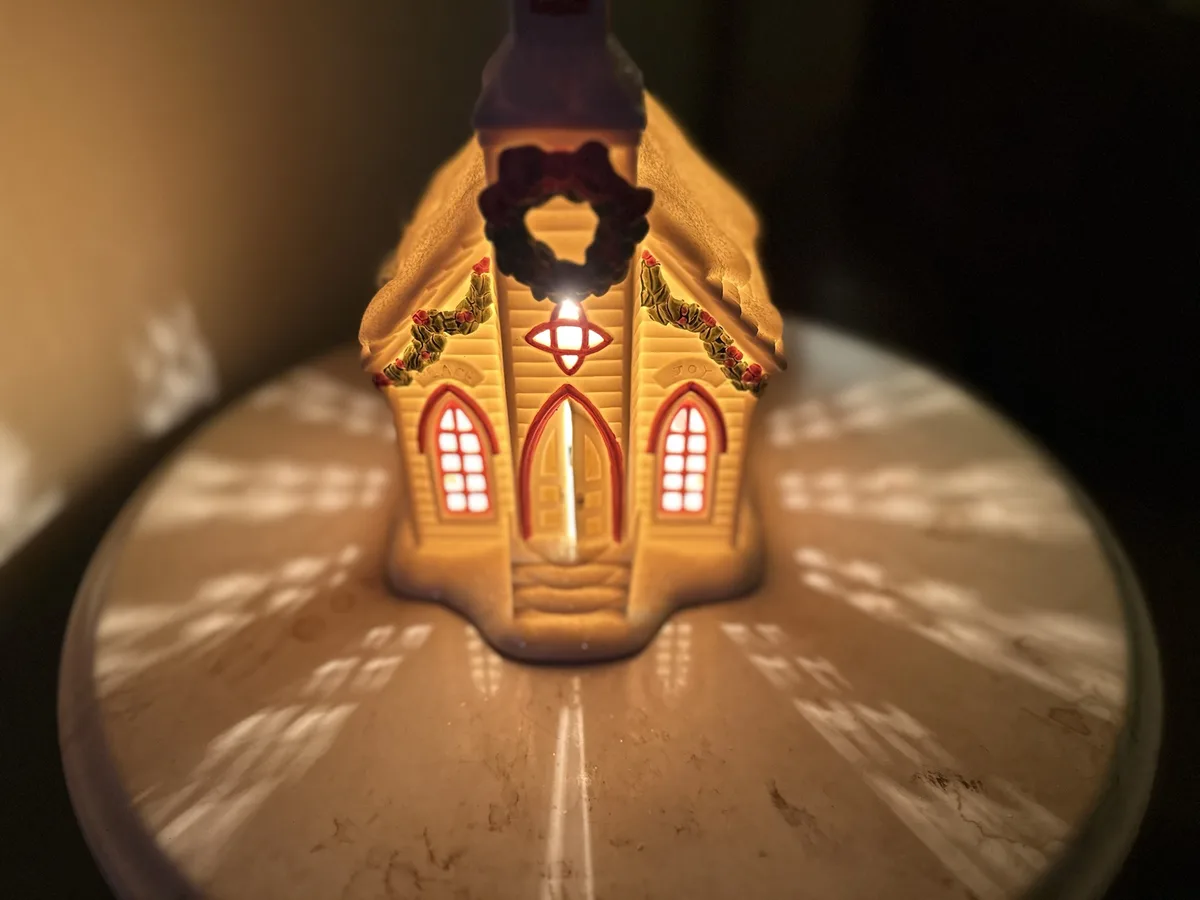 A close-up photo of a lit, yellow plastic Christmas church ornament with red-trimmed windows and a wreath on its front…