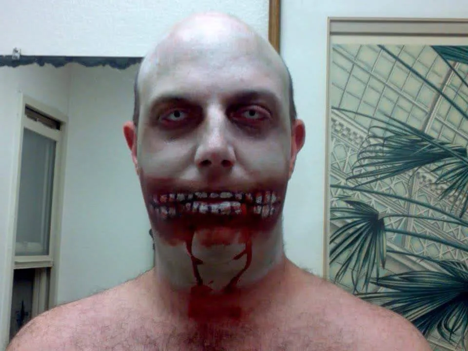 A man with a bald head and pale makeup stares directly at the camera, his mouth grotesquely altered to appear as if his…
