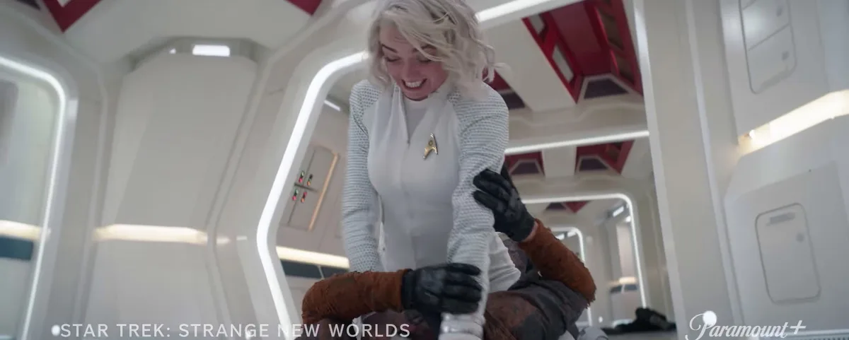 A woman in a Star Trek uniform with a gold insignia smiles while being held by someone in brown, muddy clothing inside a…