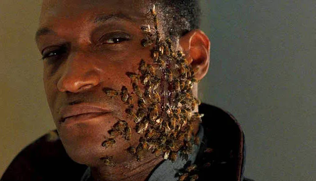 A close-up of a man with a calm, slightly amused expression as dozens of bees crawl over his face and neck. The bees are…