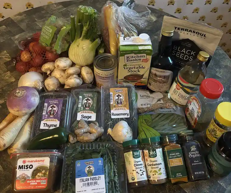 A table is covered with a large assortment of fresh produce, spices, and pantry staples including mushrooms, onions, garlic…