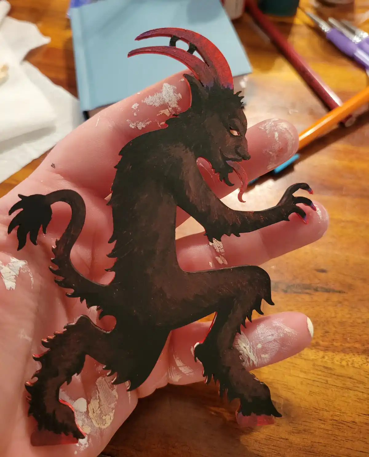 A hand holds a small, hand-painted cutout of a demonic creature with horns, claws, and a tail, showing paint smudges on the…