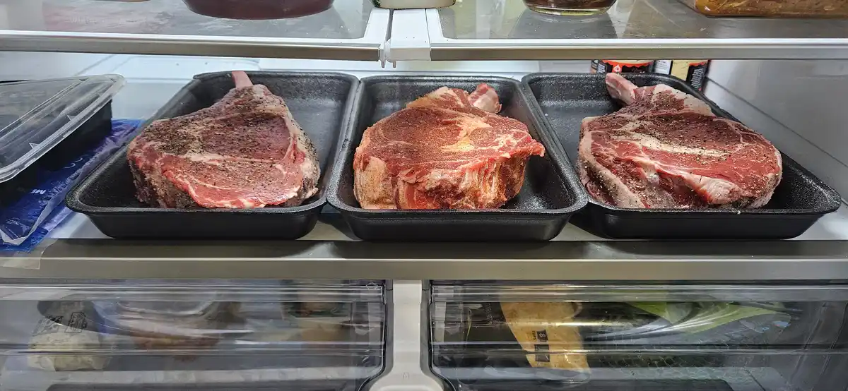 Three large, raw, seasoned beef steaks sit in black trays on a refrigerator shelf, with the overlaid text reading 'i bought…