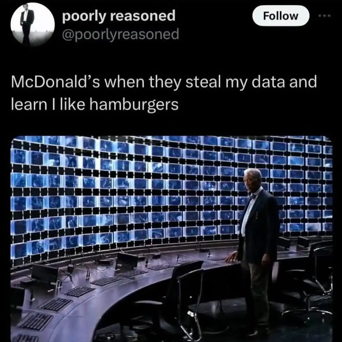 A man in a suit stands in a high-tech control room with a wall of glowing blue monitors behind him. The overlaid text reads…