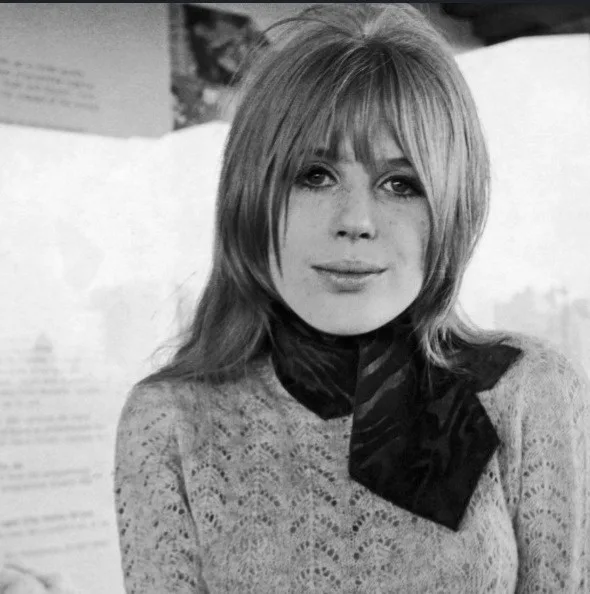 A black-and-white photo of a woman with shoulder-length hair and bangs, wearing a patterned sweater with a dark bow at the…