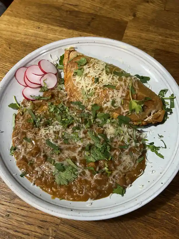 A plate of food featuring a large, folded, golden-brown pastry or empanada resting on a bed of brown bean stew, both topped…