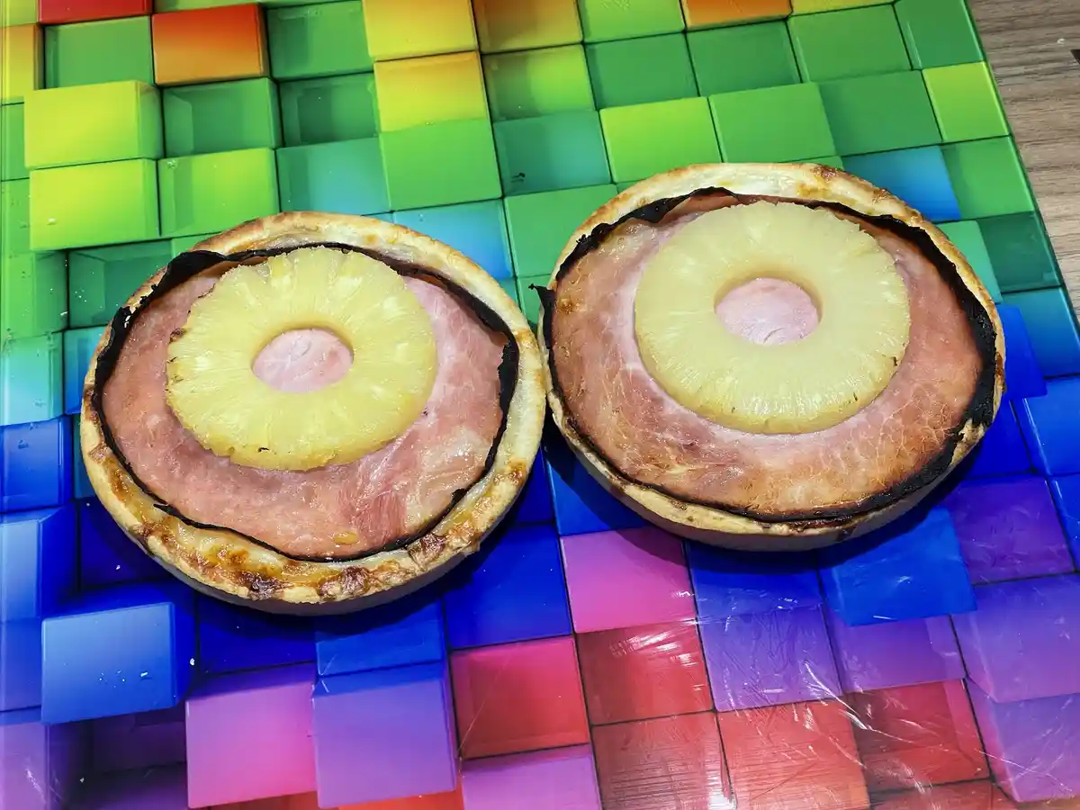 Two halves of a ham and pineapple pie sit on a colorful rainbow cube background, each half showing a slice of pineapple atop…