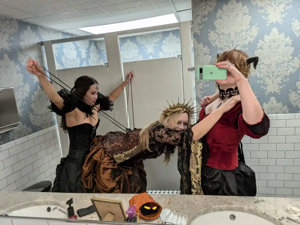 Three women in elaborate gothic or fantasy costumes are posing in a public restroom mirror. One woman in a black dress pulls…