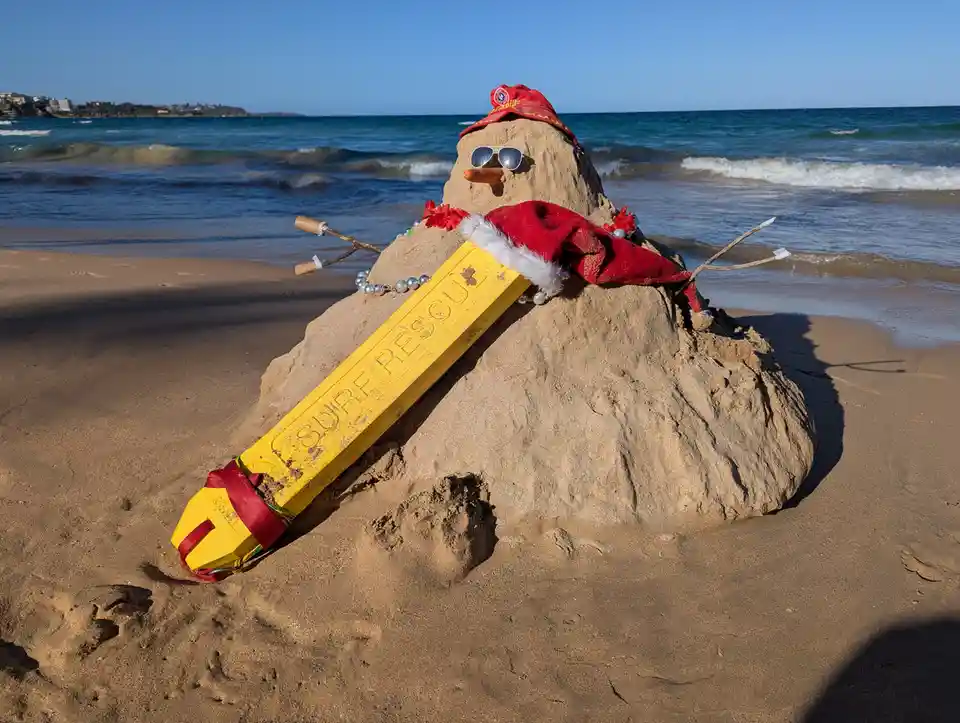 A sand sculpture on a sunny beach, shaped like a snowman, wears a red Santa hat, sunglasses, and a red scarf. It holds a…