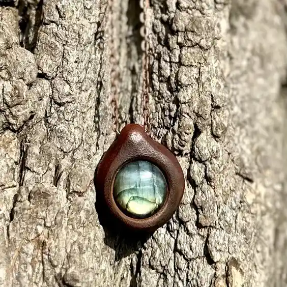A close-up photo shows a copper-colored necklace with a teardrop-shaped wooden pendant holding a labradorite stone, hanging…