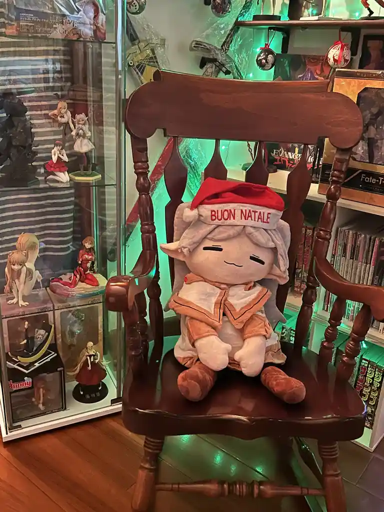 A plush toy with elf-like ears and a Santa hat that reads 'BUON NATALE' sits in a wooden rocking chair. Behind it, a glass…