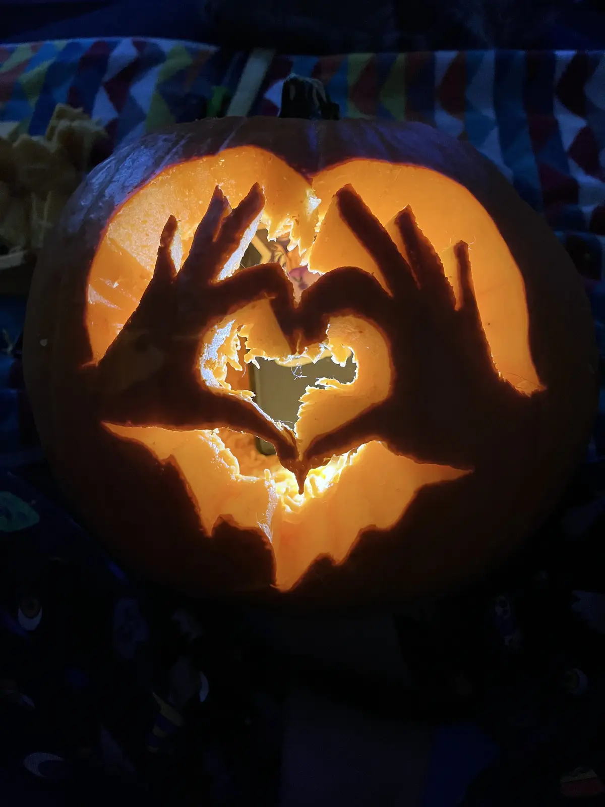 A close-up of a carved Halloween pumpkin illuminated from within, featuring two hands cut into the pumpkin's face forming a…