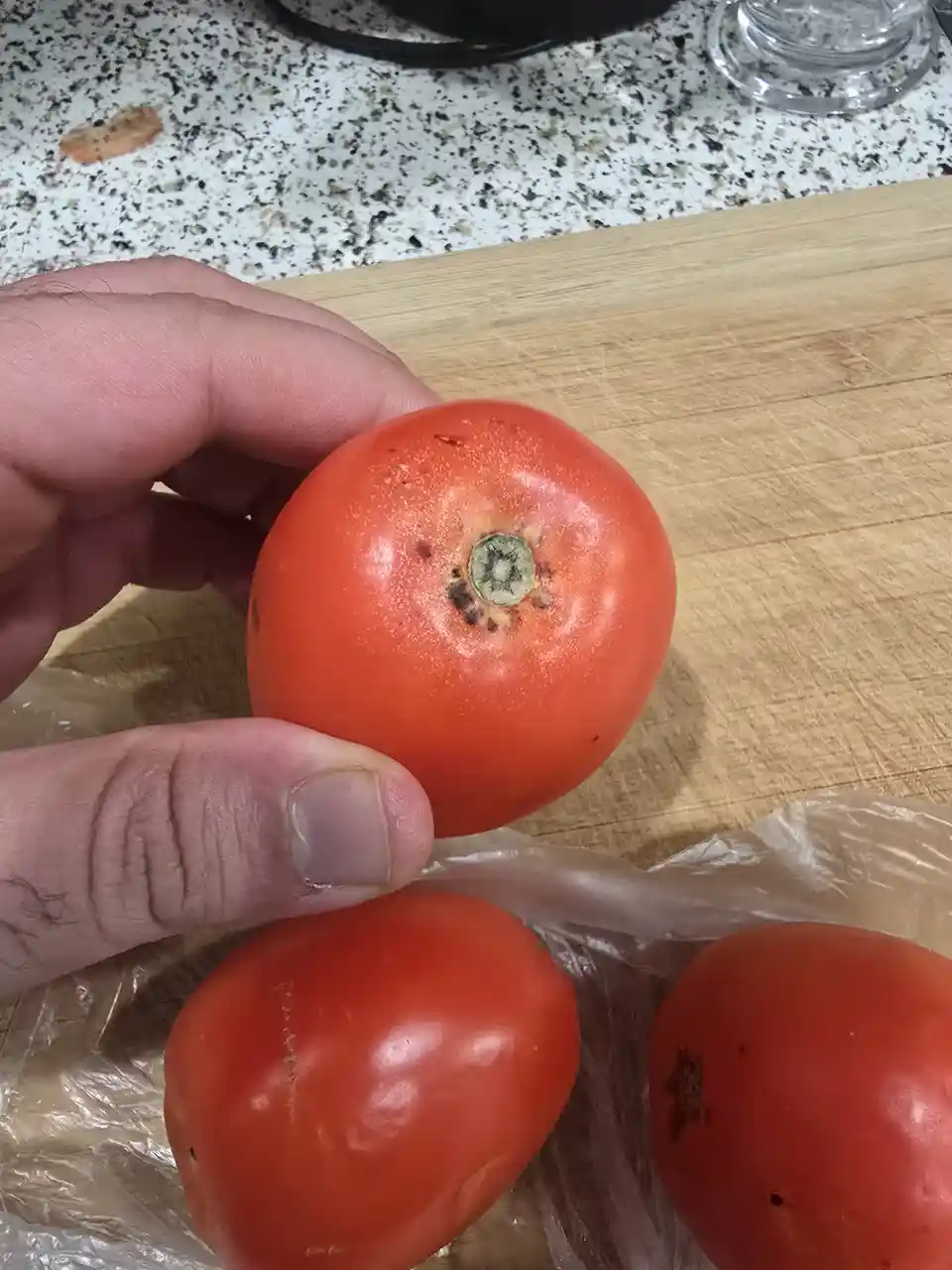 A hand holds a red tomato with a dark, rotten-looking stem area, while two other tomatoes sit in a plastic bag on a wooden…