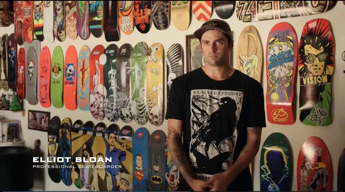 A man identified as Elliot Sloan, a professional skateboarder, stands in front of a wall covered with numerous colorful…