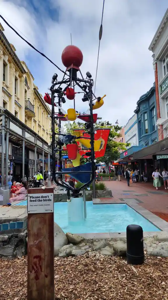 A public fountain with colorful teapots and cups spraying water is surrounded by pedestrians in a city street. A sign next…