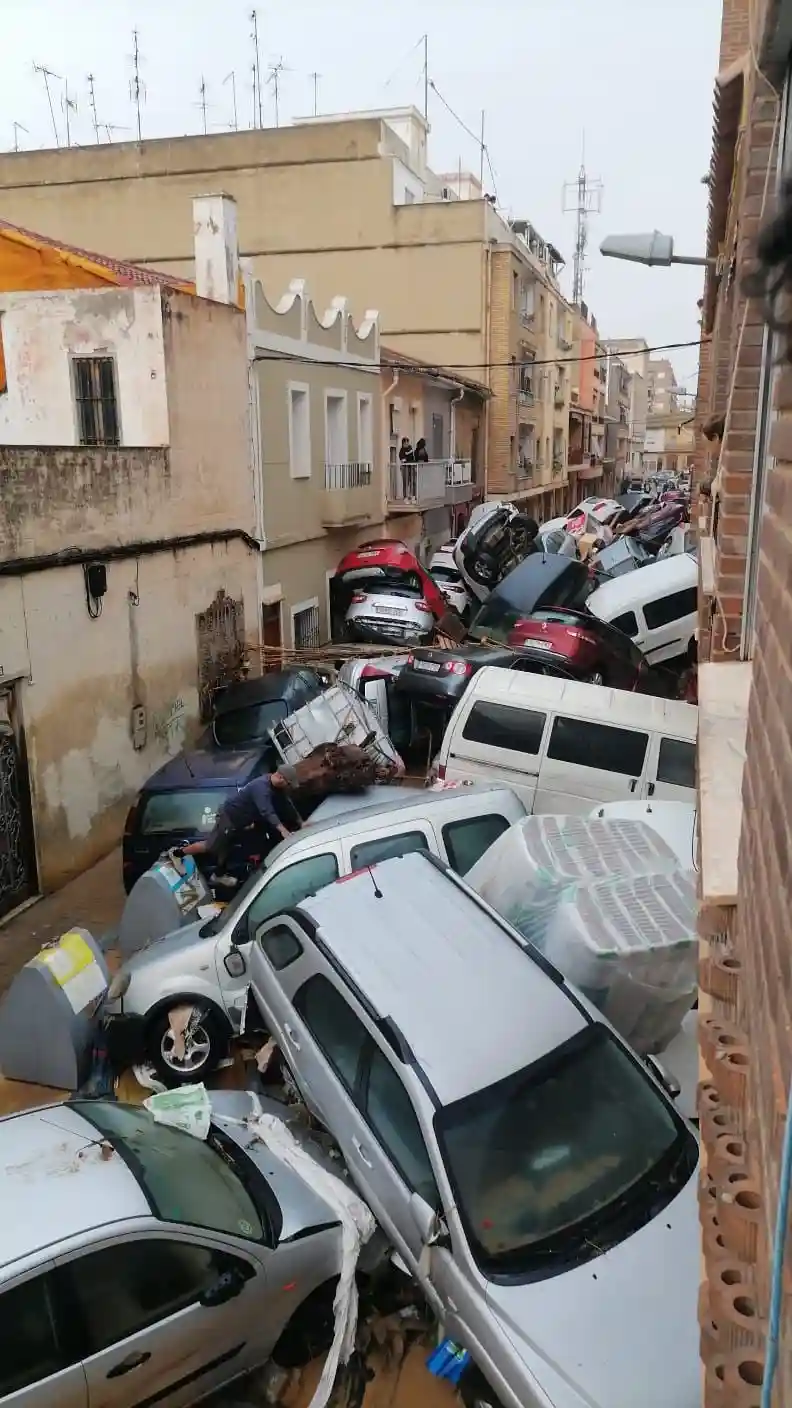 A narrow urban street is completely blocked by a chaotic pile of wrecked and crushed cars, some with hoods open and others…