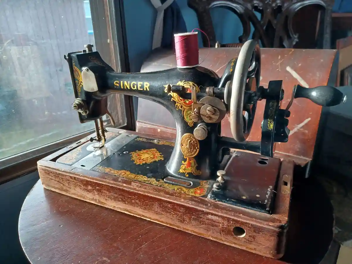 An antique black Singer sewing machine with ornate gold and red detailing sits on a wooden table near a window, topped with…