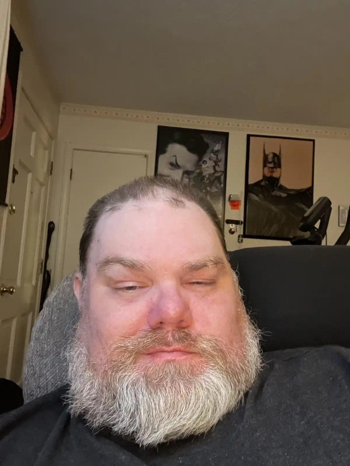 A man with a full gray beard and closed eyes sits in a chair, looking relaxed or tired. Behind him on the wall are two…