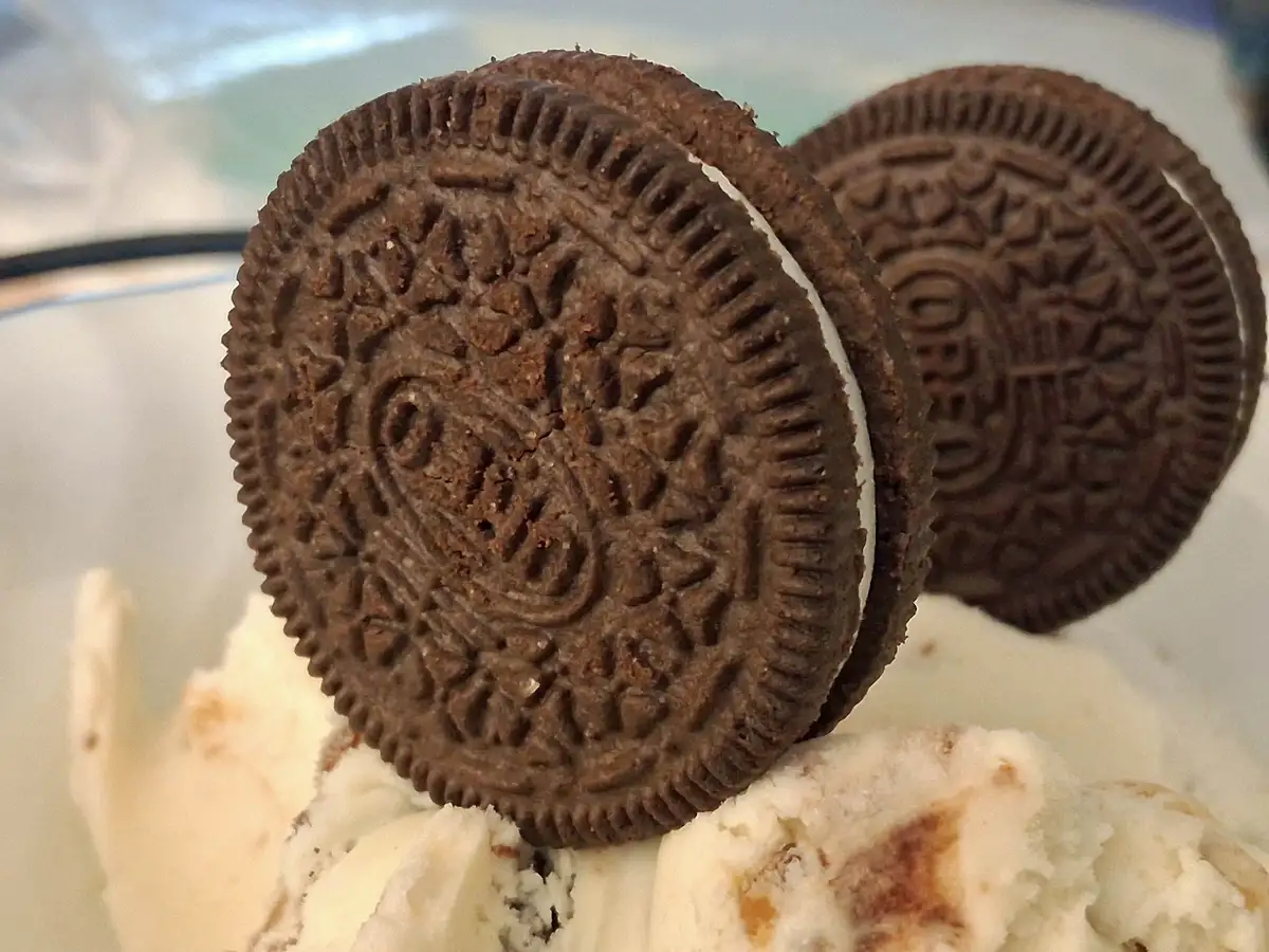 A close-up photo shows two chocolate sandwich cookies with white cream filling, standing upright in a bowl of vanilla ice…