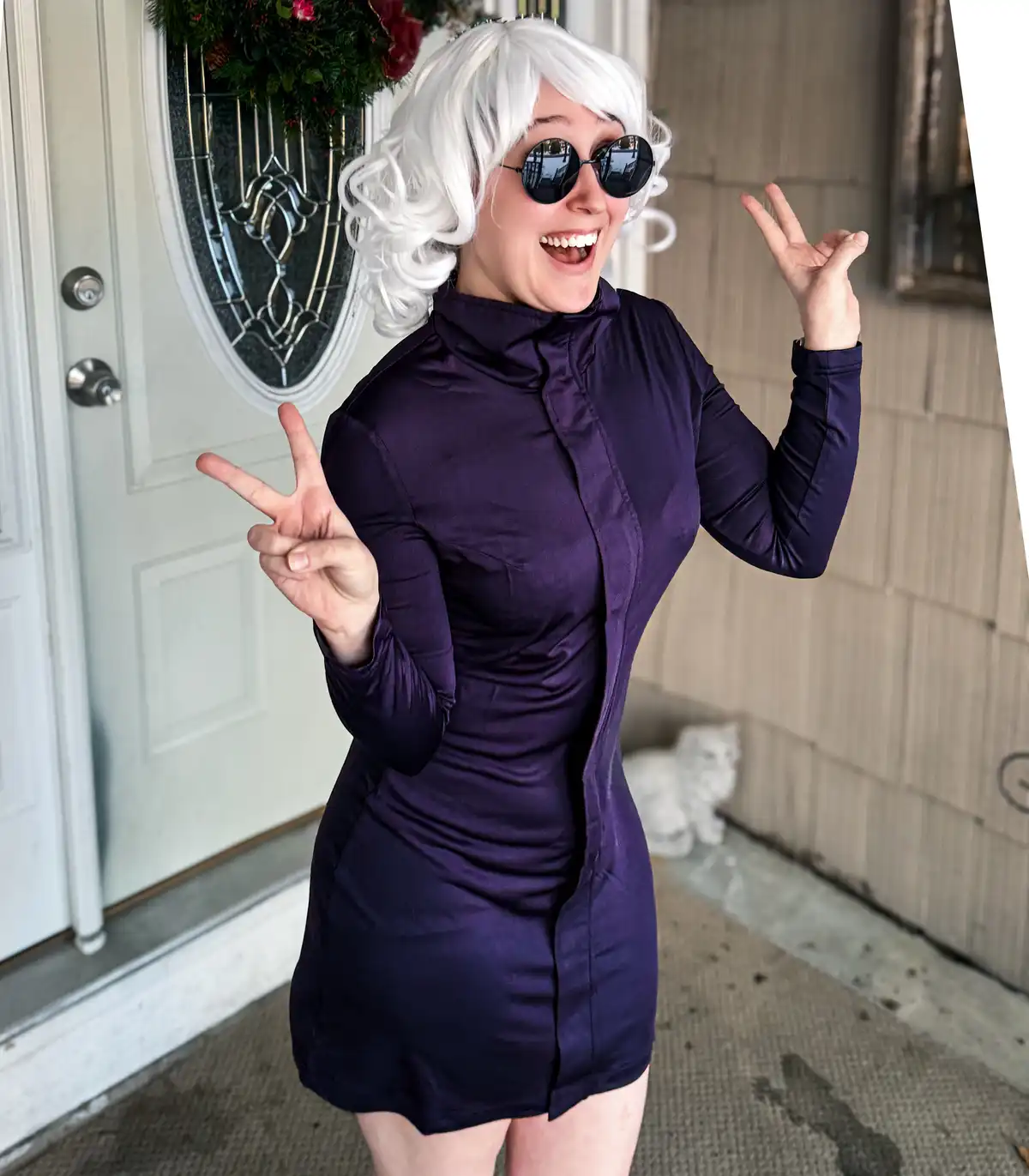 A person with a white wig and dark sunglasses poses cheerfully in front of a white door, wearing a tight-fitting purple…