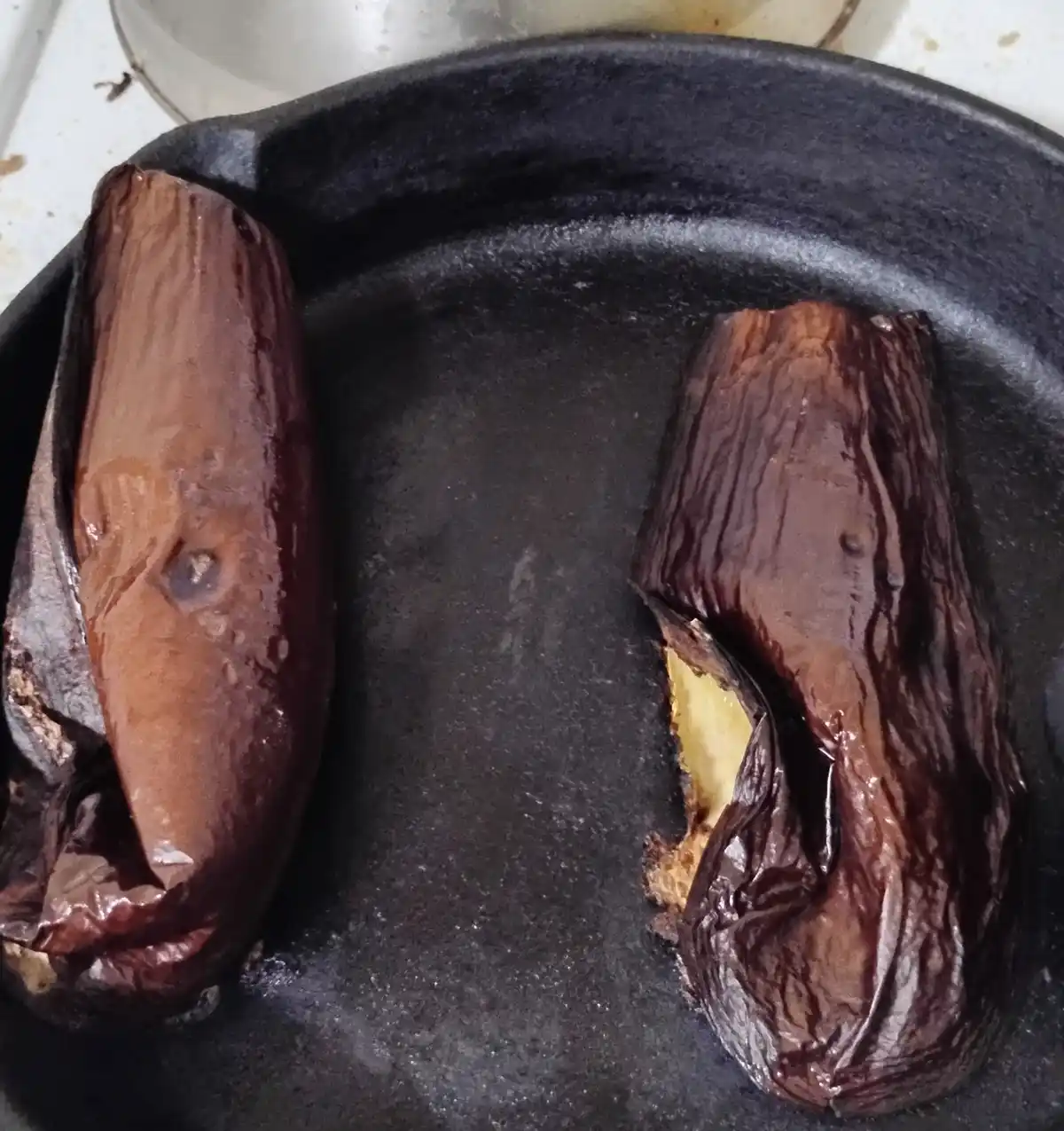 Two roasted eggplants sit in a black cast iron skillet; one is whole with wrinkled dark skin, the other is sliced open to…