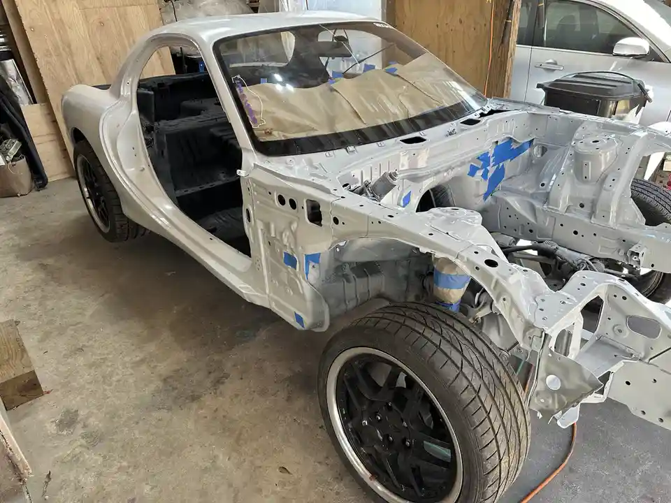 A silver car body is shown in a garage, stripped of its interior and most panels, with blue painter's tape on exposed metal…