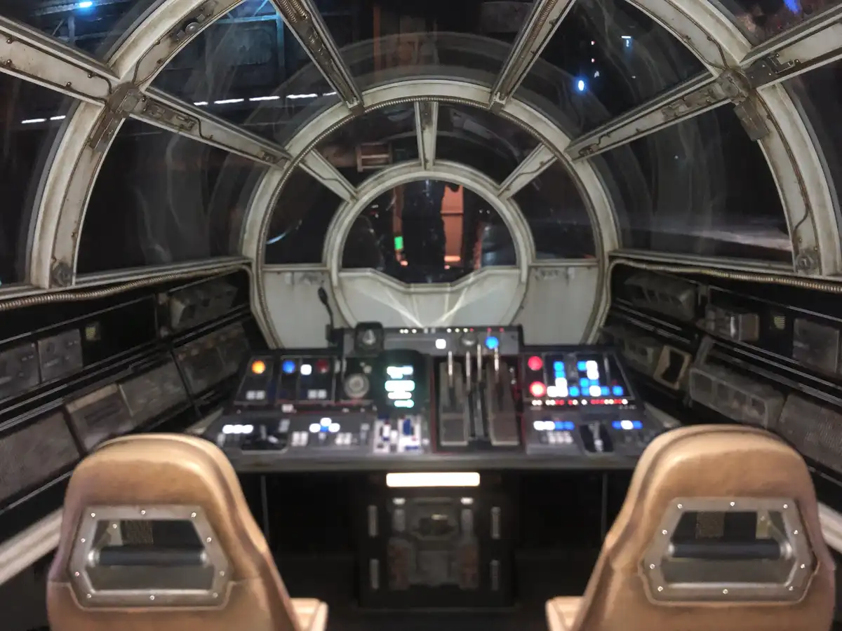 The image shows the cockpit of the Millennium Falcon from Star Wars, featuring two pilot seats, a complex control panel with…